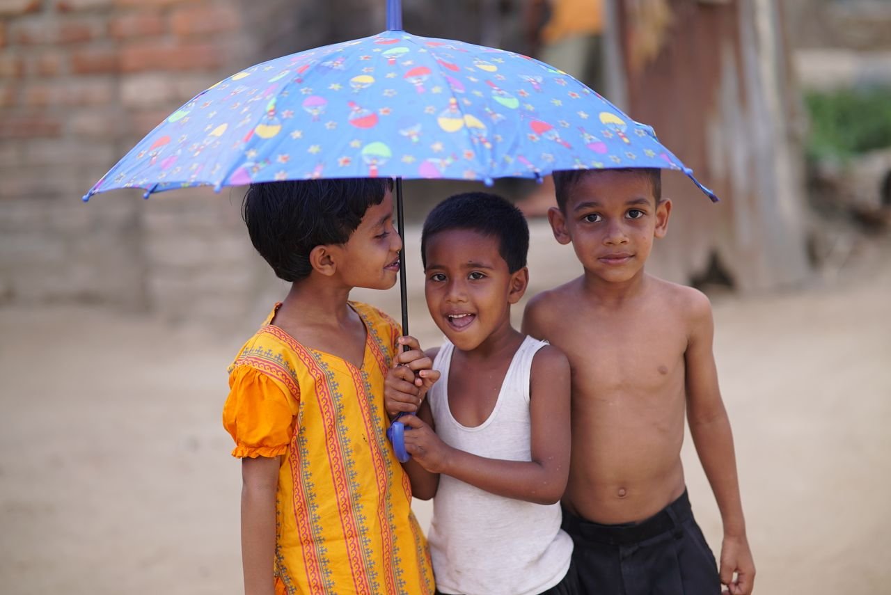 Three smiling children play under a vibrant blue umbrella, enjoying a sunny day outdoors.