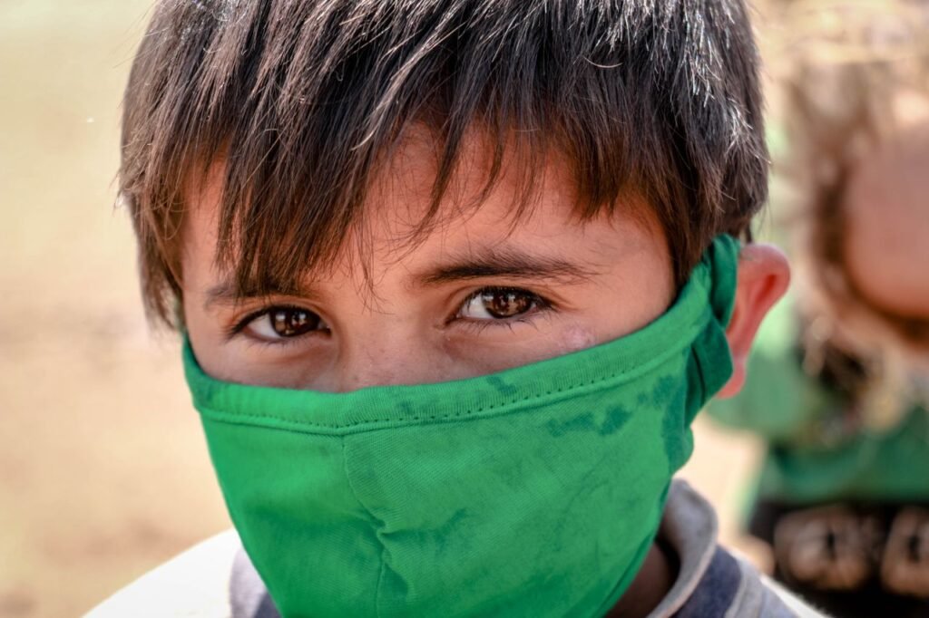 A close-up of a young boy wearing a green face mask, conveying innocence and resilience in an outdoor setting.